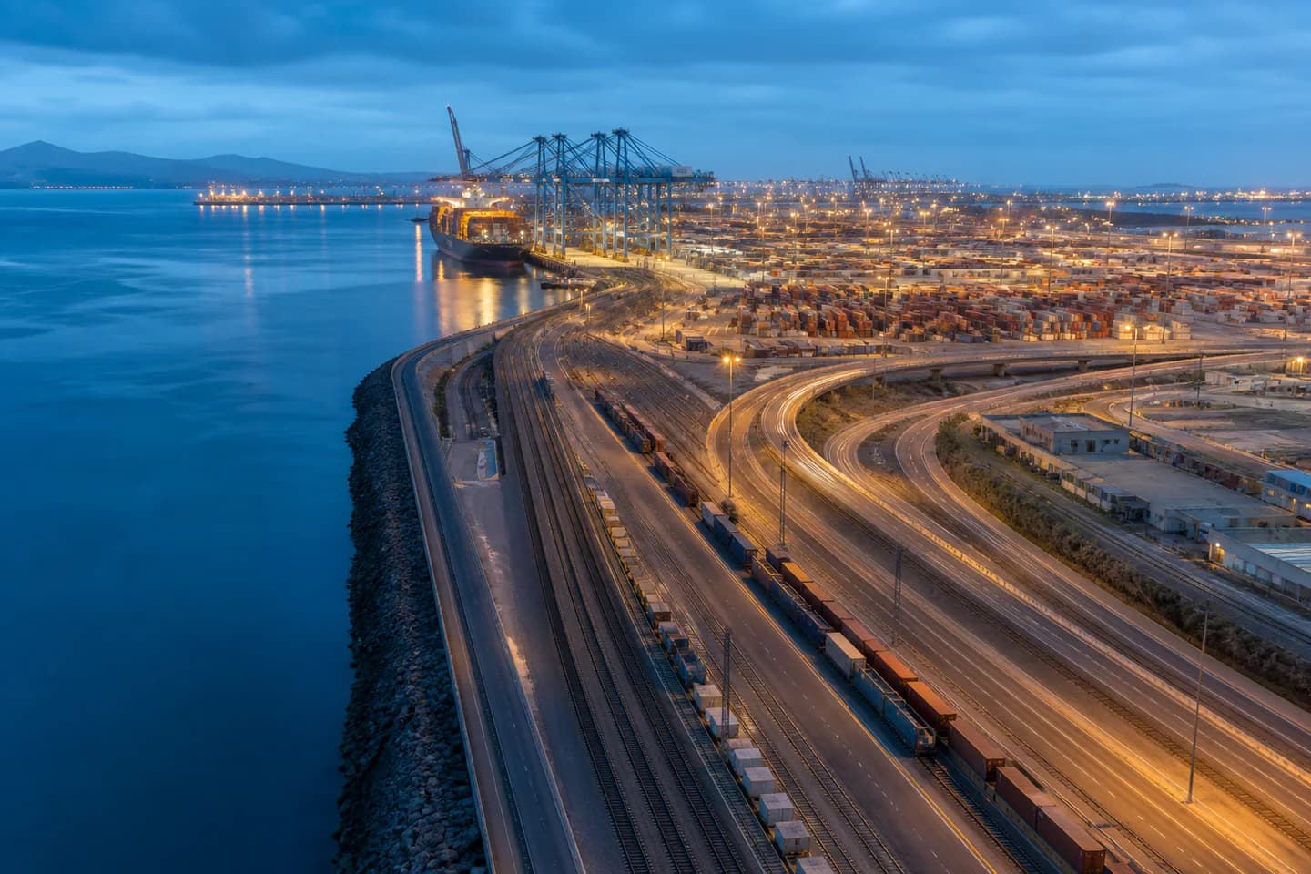 Coastal freight terminal with cargo vessel, rail lines, and roads converging at blue hour